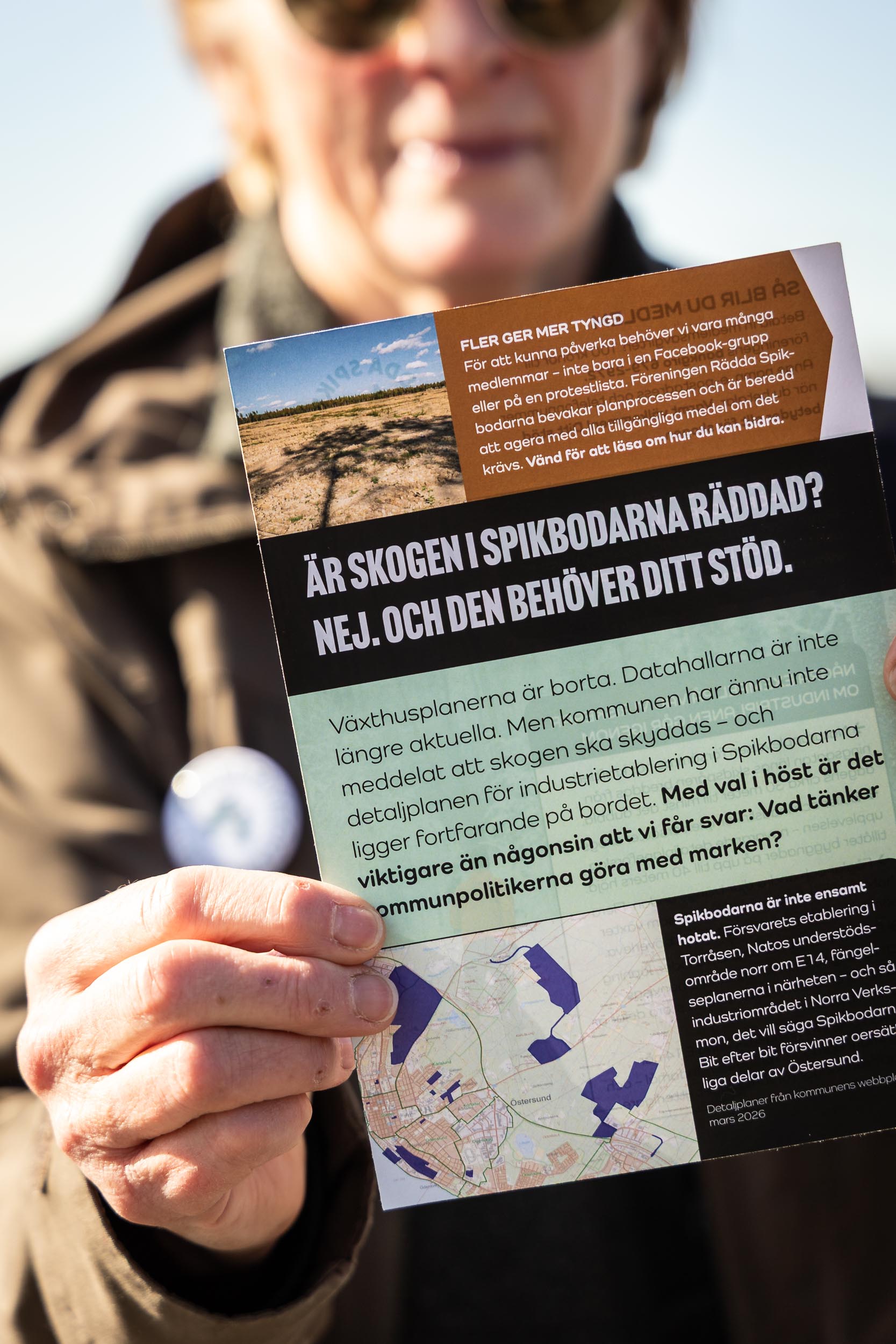 En hand håller upp en bunt broschyrer. I bakgrunden syns ett pågående kommunfullmäktige-möte i Östersund.